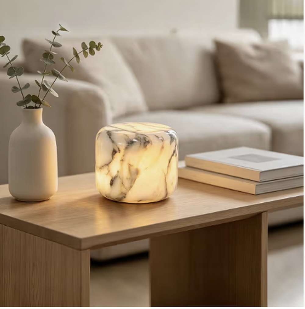 Una pequeña lámpara de mármol con luz cálida sobre una mesa de centro de madera, junto a un jarrón de eucalipto y dos libros.