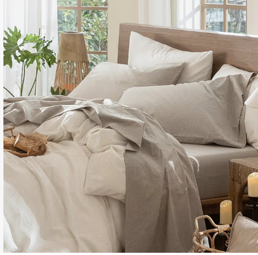 Una cama hecha con sábanas de algodón lavado en tonos avena y piedra, con cabecero de madera y una vela al lado.